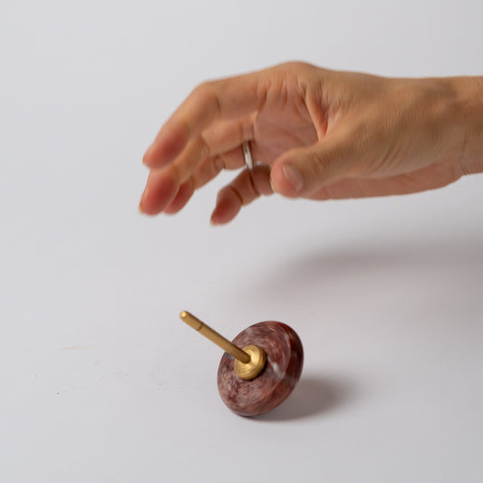 A hand suspended over a red jasper spinning top with a gold spindle, spinning at an angle against a clean white background.