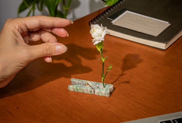 Hand reaching toward a small white flower held by a minimalist clothespin vase, carved from natural green serpentine stone, on a warm wooden desk.