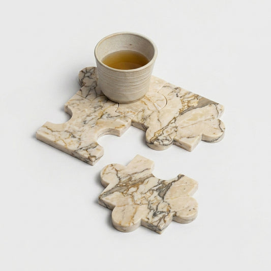 Three creamy soapstone coasters, interlocked like puzzle pieces, hold a small ceramic cup. A fourth piece rests separately below on a white background.