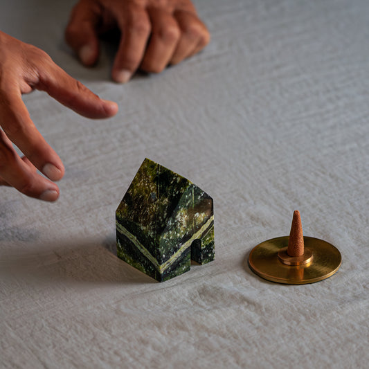 Two hands reach toward a green serpentine house with distinct light veins, resting on a fabric surface. Next to it, an incense cone sits on a round gold base.