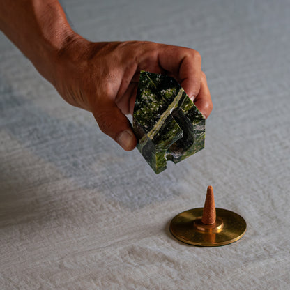 A hand lifts a small carved green serpentine house, showing its hollow interior. Below, on a light fabric, an incense cone rests on a round gold base.