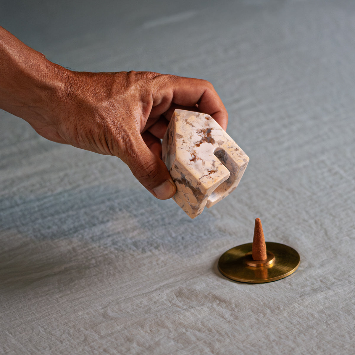 A hand lifts a small carved creamy soapstone house, showing its hollow interior. Below, on a light fabric, an incense cone rests on a round gold base.