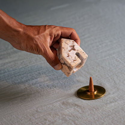 A hand lifts a small carved creamy soapstone house, showing its hollow interior. Below, on a light fabric, an incense cone rests on a round gold base.