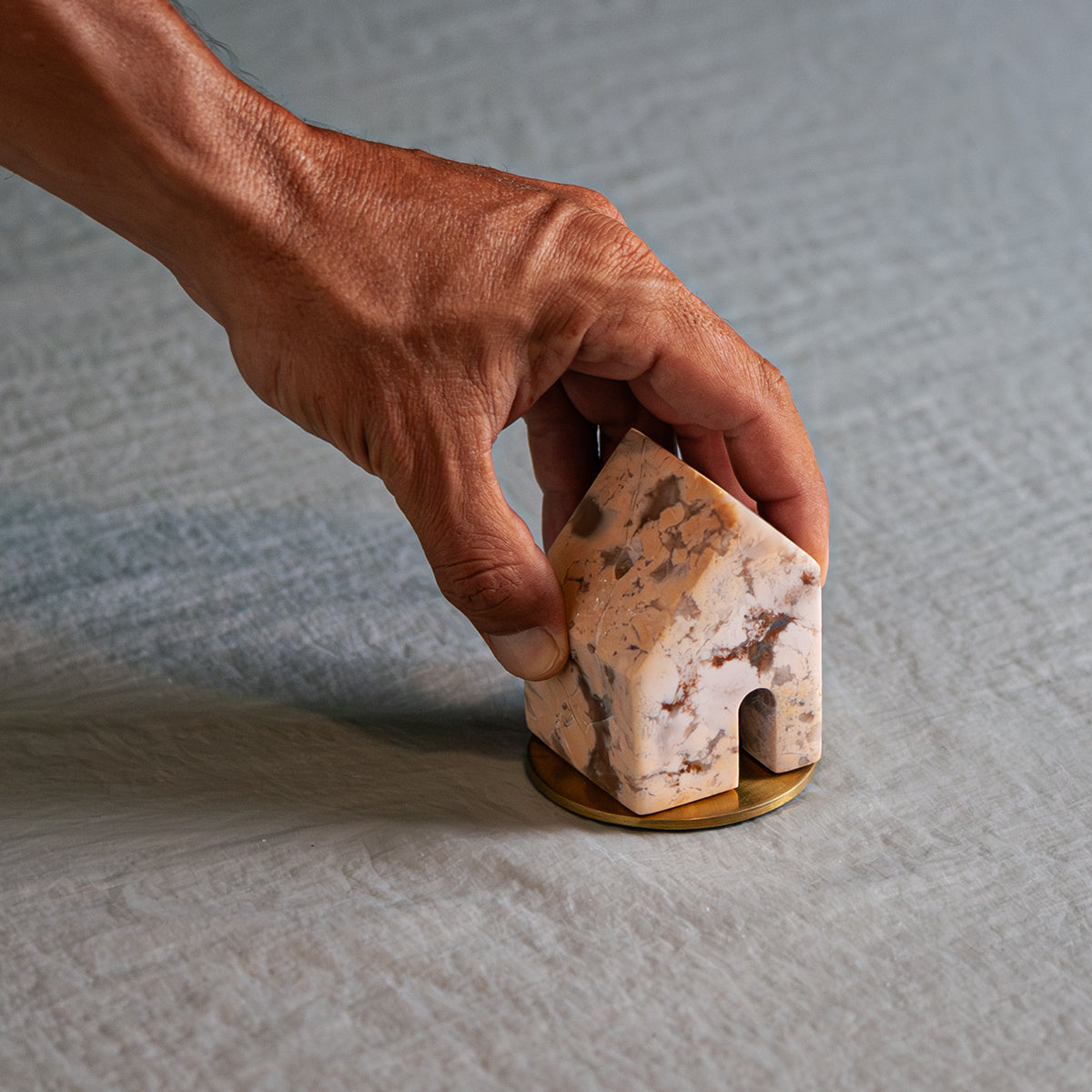 A hand holds a small carved creamy soapstone house, placing it onto a round gold base resting on a light fabric.