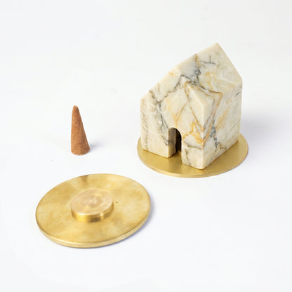Creamy soapstone house-shaped incense holder resting on a gold base. Next to it lies a loose incense cone and a second gold base against a clean white background.