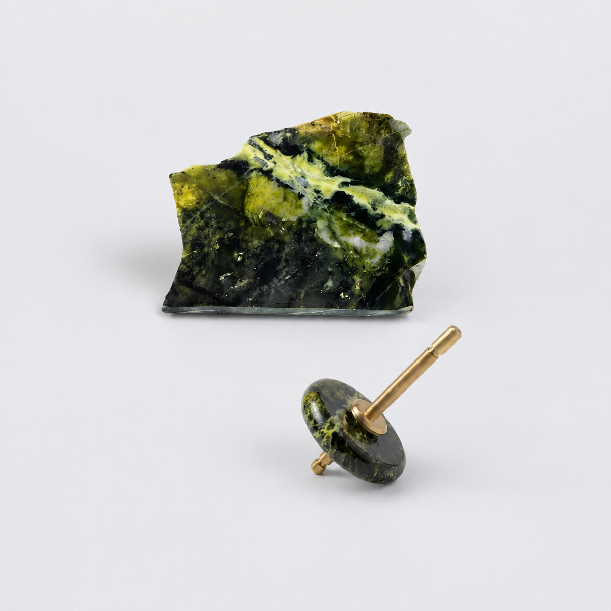 Spinning top with a polished green nephrite disc and gold spindle, resting on a white background next to a raw, unpolished block of the same stone.