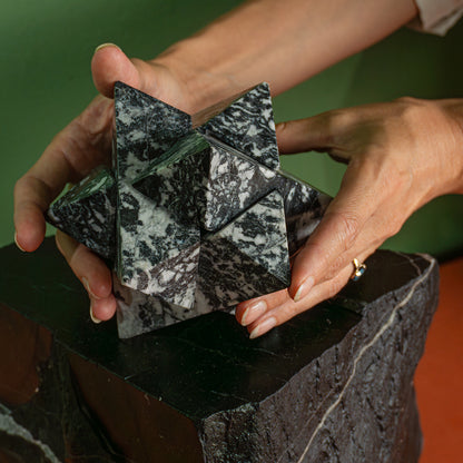 Two hands holding a large zebra jasper star, showing the scale and volume of its geometric proportions over a dark base.