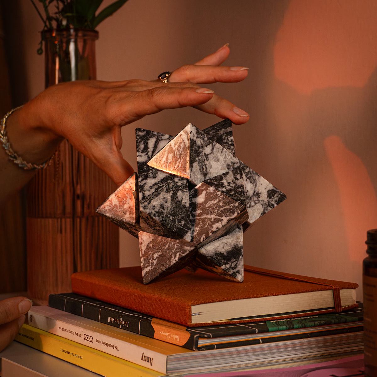 A hand interacting with the symmetry of a zebra jasper star. The three-dimensional figure with proportional cuts rests on a stack of books under warm lighting.