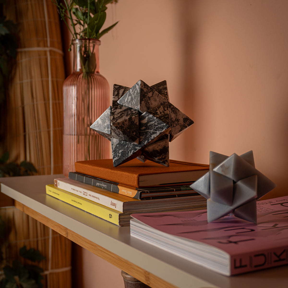 A zebra jasper star on top of books, contrasting its volume and proportions with a version carved in angelite, next to a pink vase.