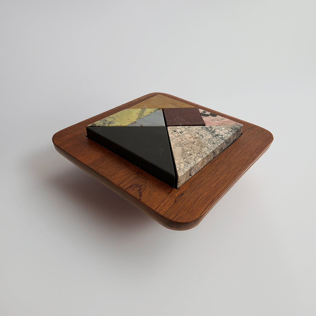 Isometric view of the puzzle assembled into a perfect square on a wooden base. The tones and textures of the natural stones stand out against a clean white background.