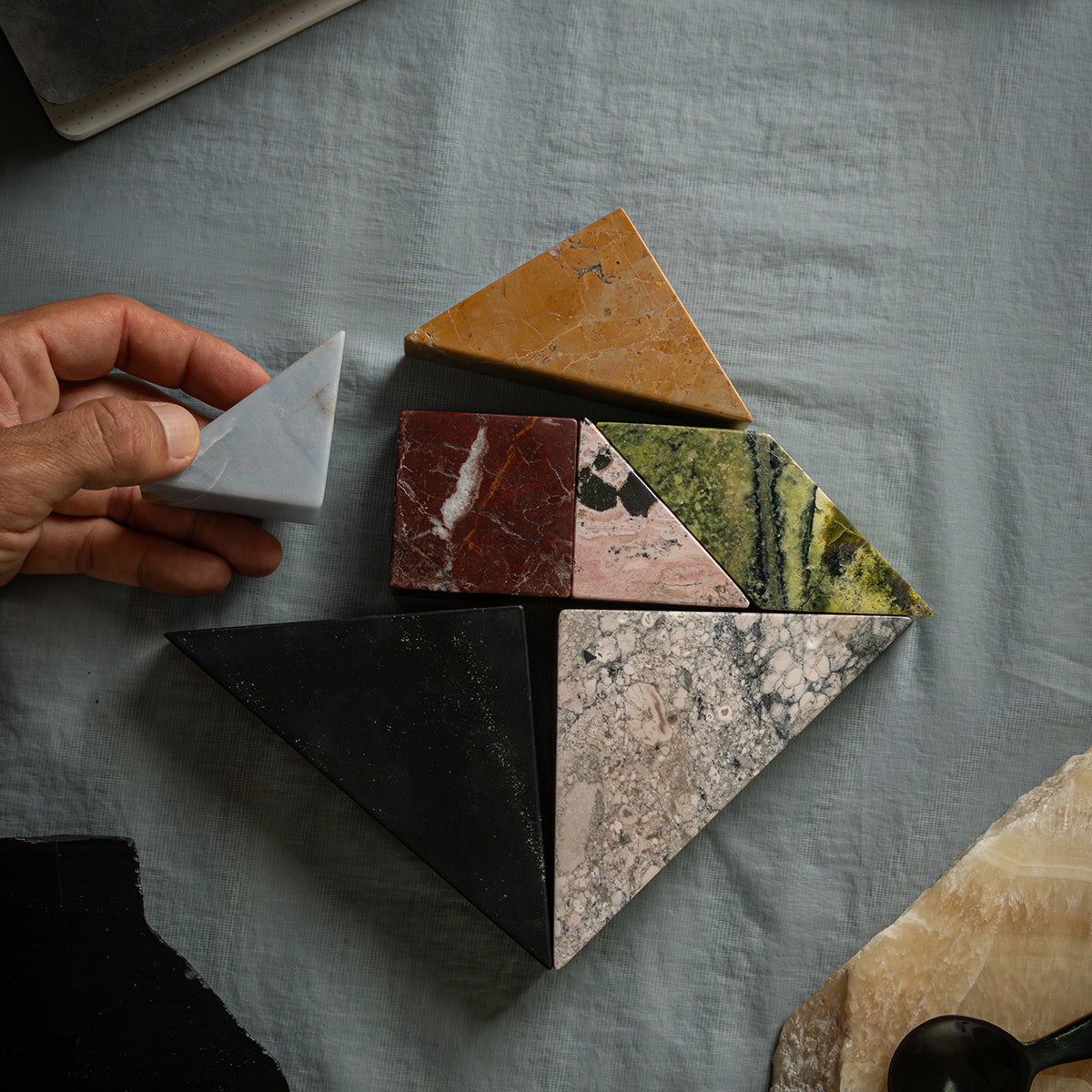 A hand placing the last triangular blue stone piece to complete the puzzle. The shot shows the real size of the pieces on the background fabric.