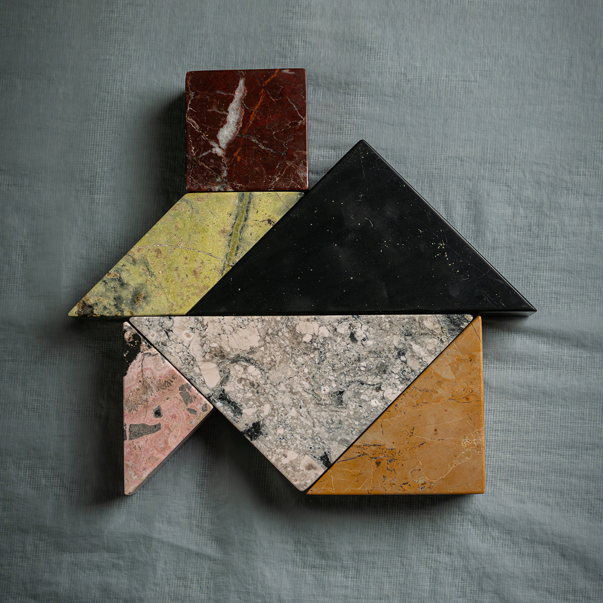 Natural stone puzzle assembled into an abstract figure. The pieces are terracotta, green, pink, and gray, resting on a grayish-blue fabric.