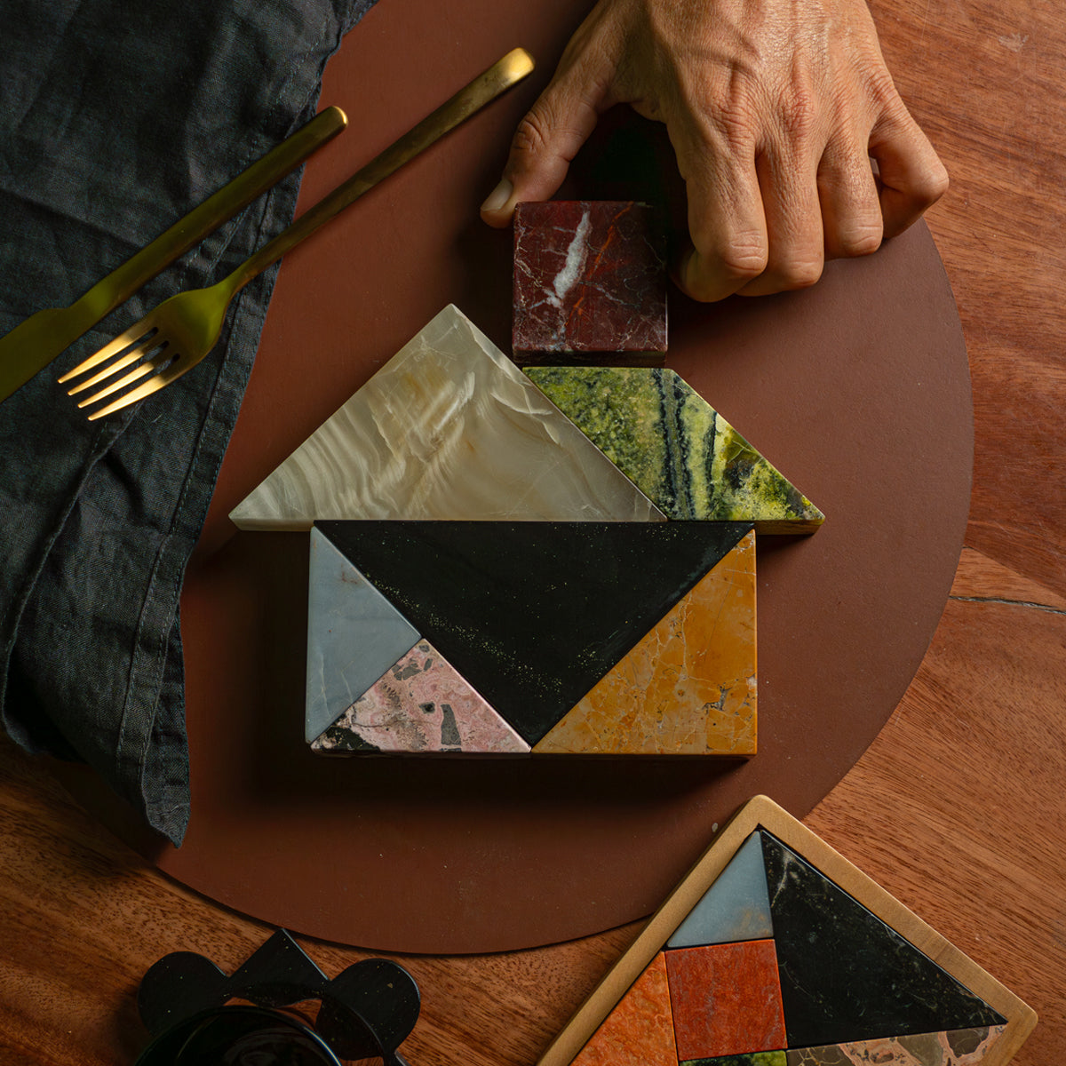 A hand placing a square reddish stone piece to complete a house-shaped tangram. The different colored pieces rest on a round brown placemat on a wooden table, next to a dark napkin and gold cutlery.