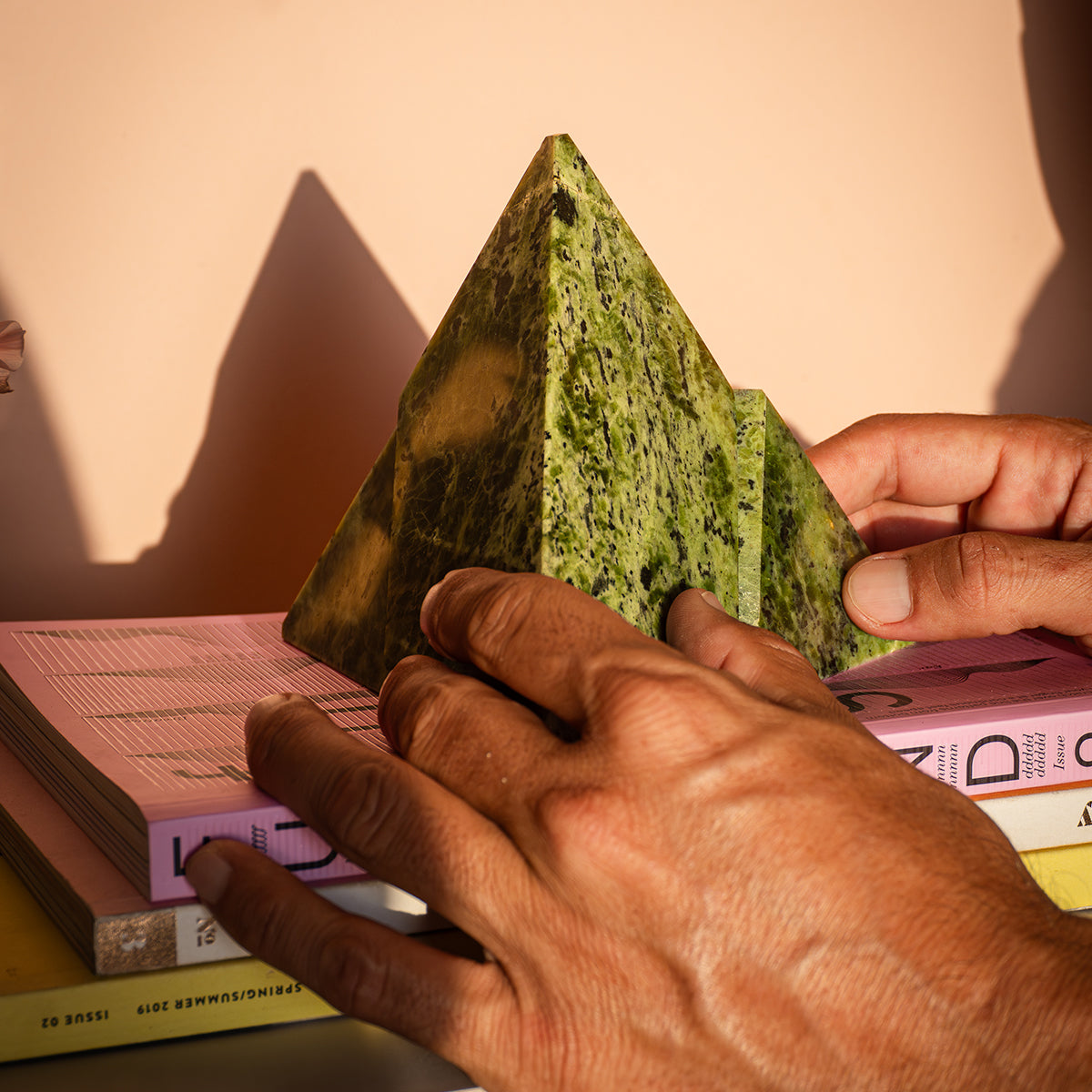 A deep green nephrite pyramid, formed by two interlocking proportional pieces, resting on books. Next to it is a white vase with yellow flowers casting shadows on the wall.