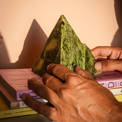 A deep green nephrite pyramid, formed by two interlocking proportional pieces, resting on books. Next to it is a white vase with yellow flowers casting shadows on the wall.