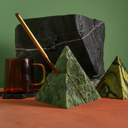 Two hands adjusting the pieces of a geometric deep green nephrite pyramid. The figure rests on a stack of books under natural light that highlights its interlocking joints.