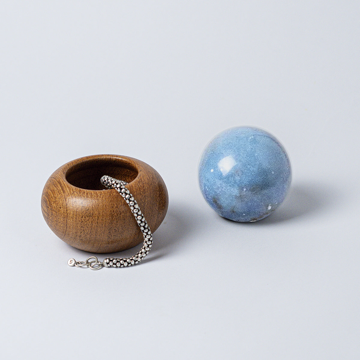 Open round wooden container with a silver chain peeking over its edge. A solid blue angelite stone handle rests next to it on a white background.