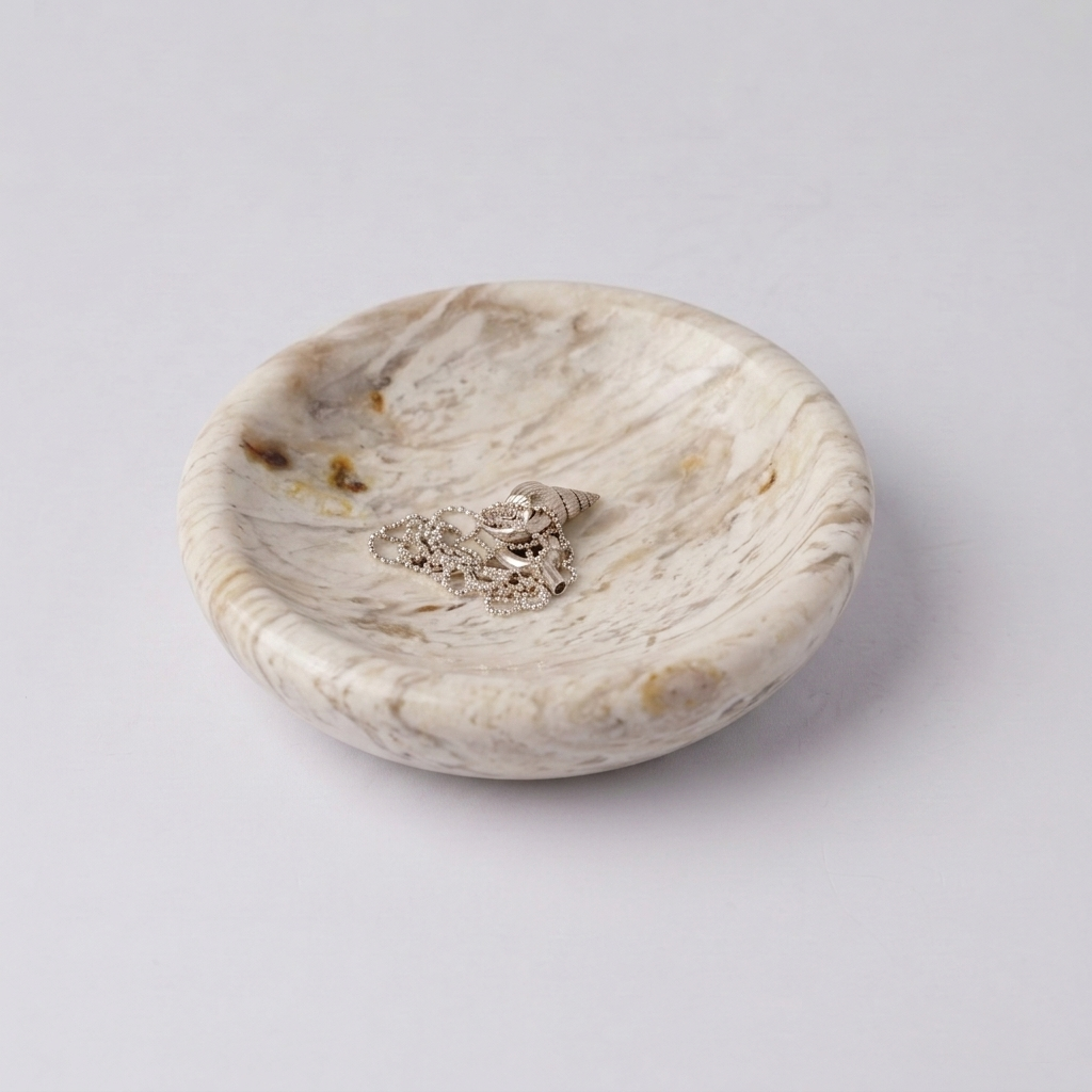 Round dish made of naturally veined creamy soapstone. Inside rests a delicate silver chain with a seashell pendant, against a white background.