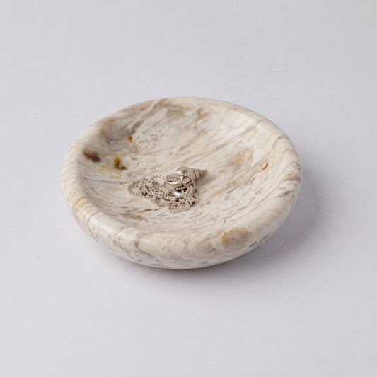 Round dish made of naturally veined creamy soapstone. Inside rests a delicate silver chain with a seashell pendant, against a white background.