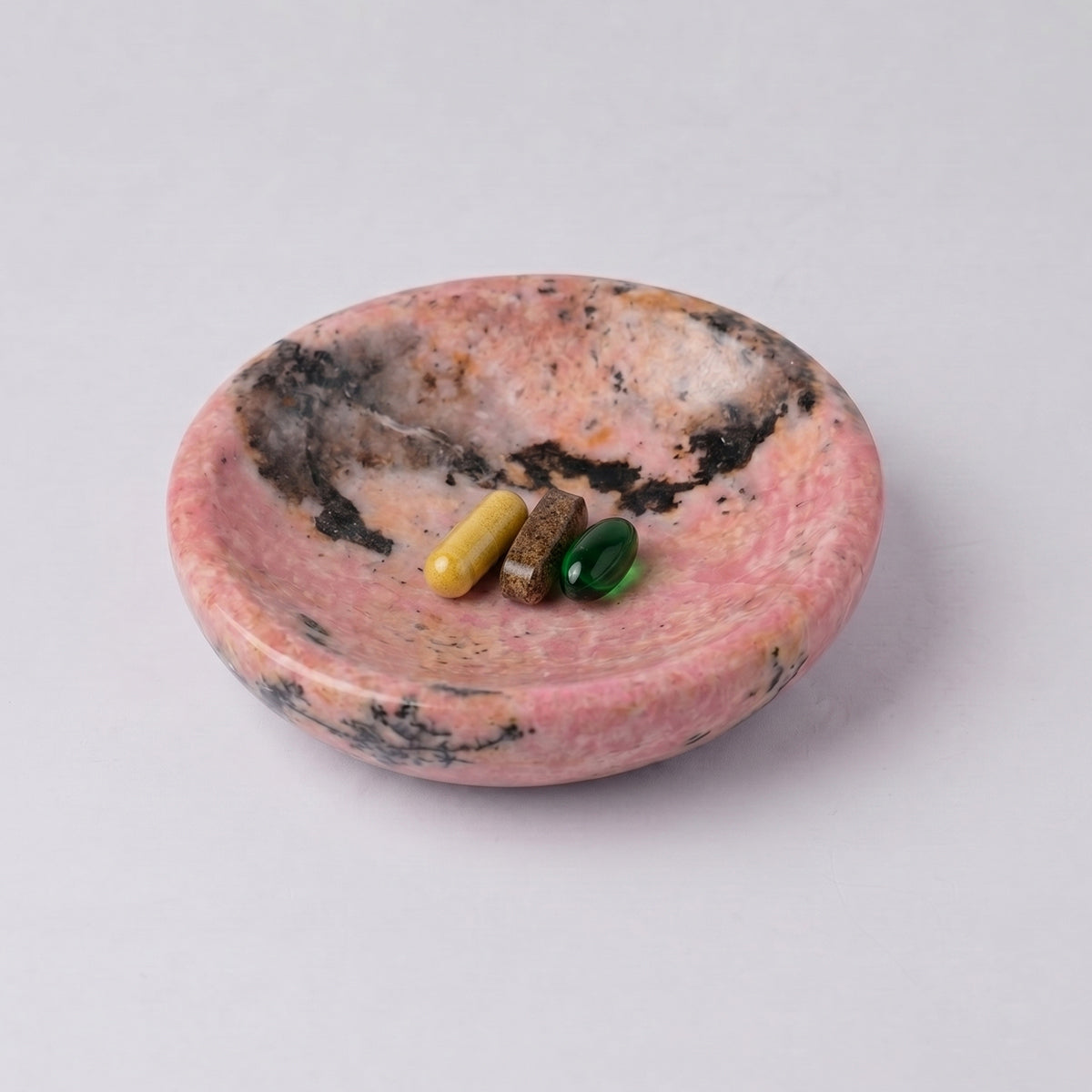 Round pink rhodonite dish with natural dark inclusions. Inside are three differently colored pill capsules (yellow, brown, and green), against a white background.