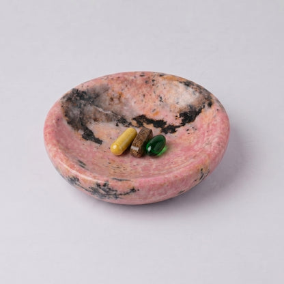Round pink rhodonite dish with natural dark inclusions. Inside are three differently colored pill capsules (yellow, brown, and green), against a white background.