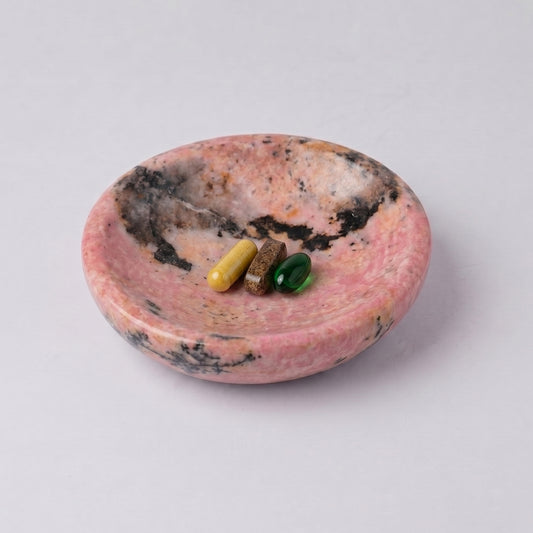 Round pink rhodonite dish with natural dark inclusions. Inside are three differently colored pill capsules (yellow, brown, and green), against a white background.