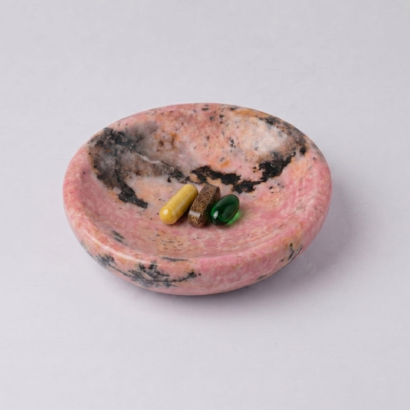 Round pink rhodonite dish with natural dark inclusions. Inside are three differently colored pill capsules (yellow, brown, and green), against a white background.