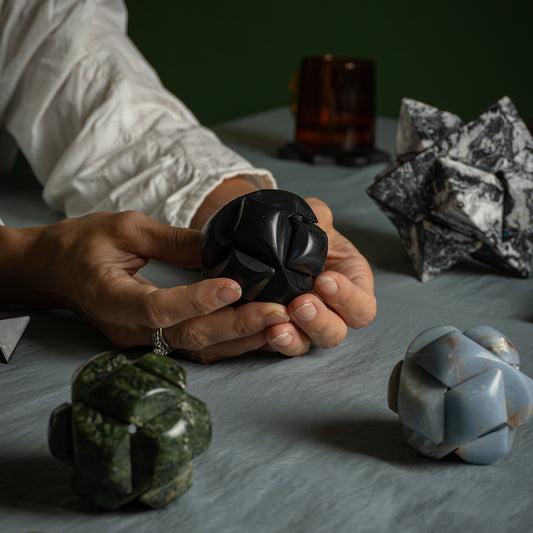 Two hands display the geometric assembly of a spherical black stone puzzle. In the foreground, the details and proportions of two other pieces in green serpentine and blue angelite can be seen.