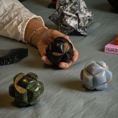 A hand holds a spherical black stone puzzle made of interlocking pieces. Resting on the fabric-covered table are similar versions carved in green serpentine and blue angelite.