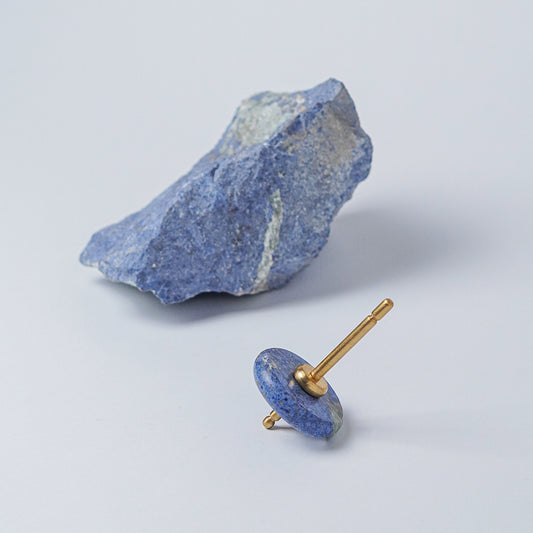 Spinning top with a polished blue dumortierite disc and gold spindle, resting on a white background next to a raw block of the same stone.