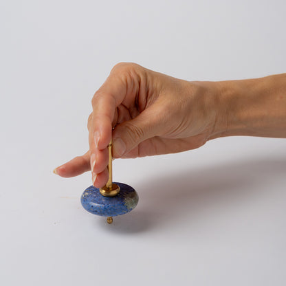 A hand holds the gold spindle of a blue dumortierite spinning top, getting ready to spin it against a clean white background.