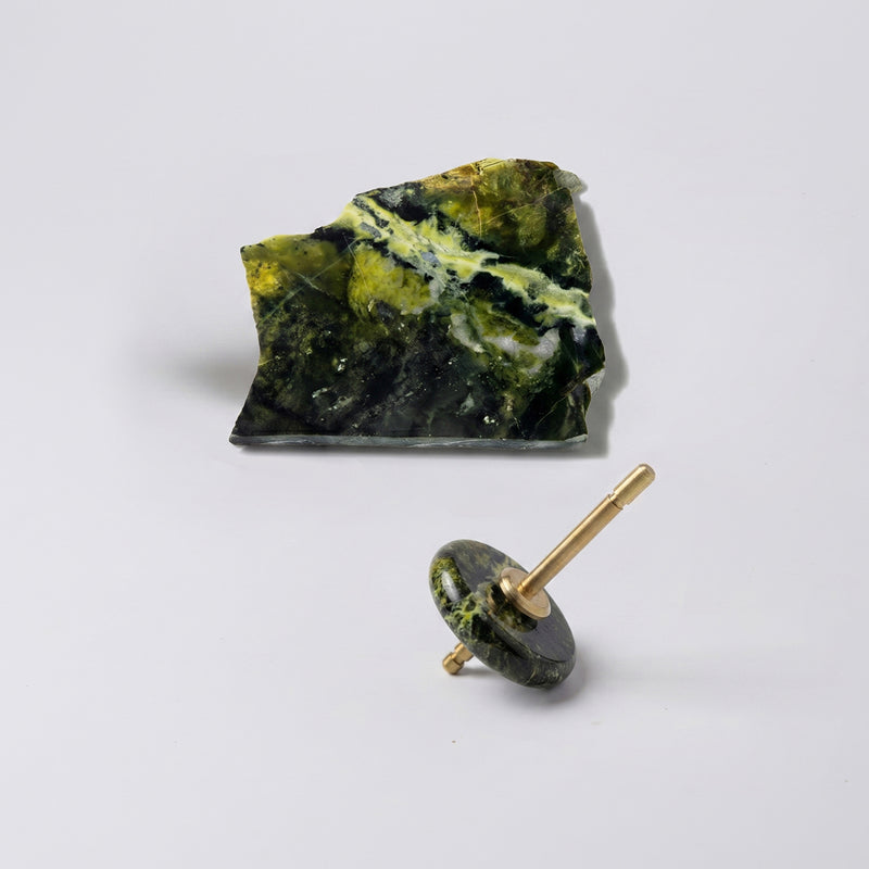 Spinning top with a polished green nephrite disc and gold spindle, resting on a white background next to a raw, unpolished block of the same stone.