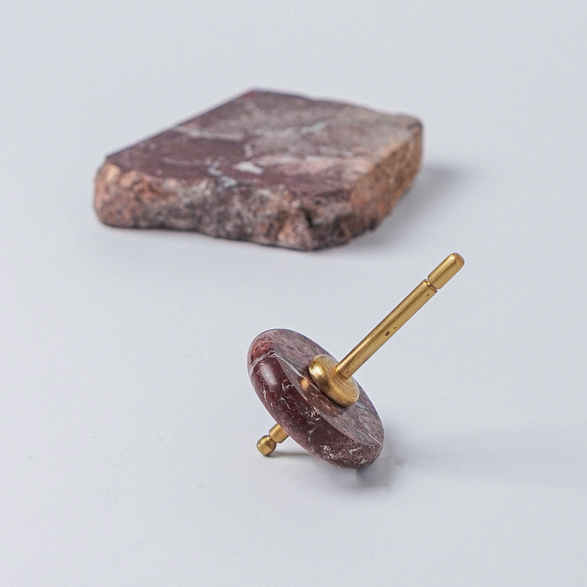 Spinning top with a polished red jasper disc and gold spindle, resting on a white background next to a raw block of the same stone.