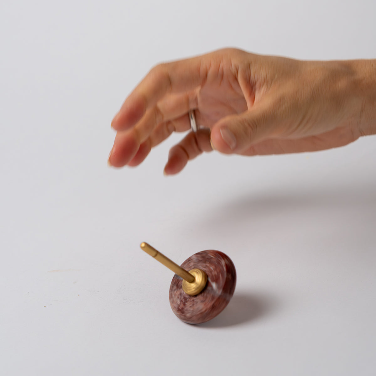 A hand suspended over a red jasper spinning top with a gold spindle, spinning at an angle against a clean white background.